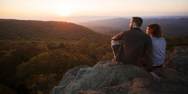 Two serene-looking people sit side-by-side on large rocks overlooking the hills and forests of Shenandoah National Park in Virginia, USA at sunset. The glow of the sun reflects on their faces.
